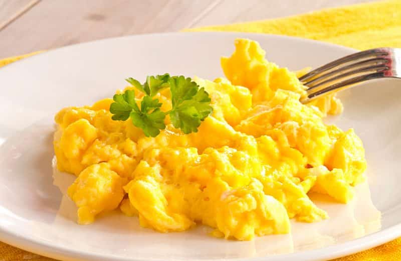 White plate with scrambled eggs on top, fork laid on eggs, on top of yellow placemat on wooden table