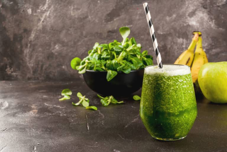 Green nutritional shake with straw sitting in front of greens, green apple, and banana