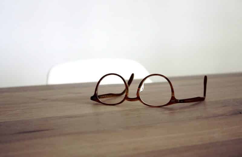 What to know before buying eyeglasses online. Image is of pair of simple brown circular lens eyeglasses sitting upside down on wooden desk.