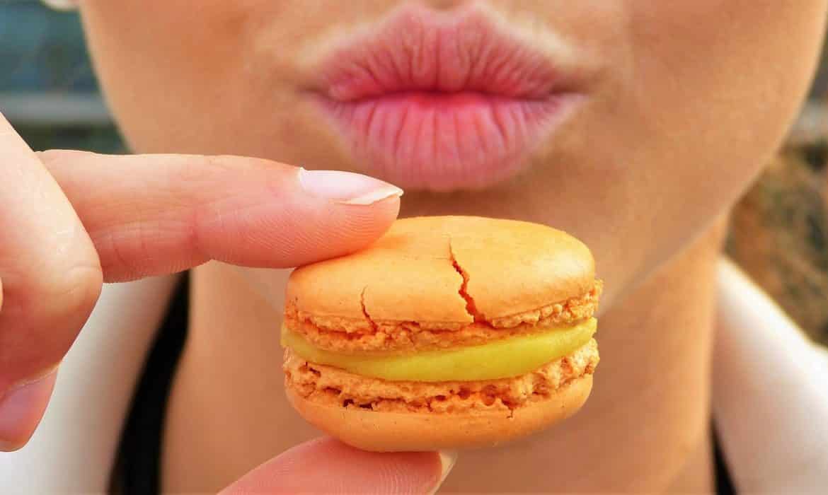 Outsmart your appetite – close up shot of woman’s pursed lips as she holds orange macaron cookie in front of her face.