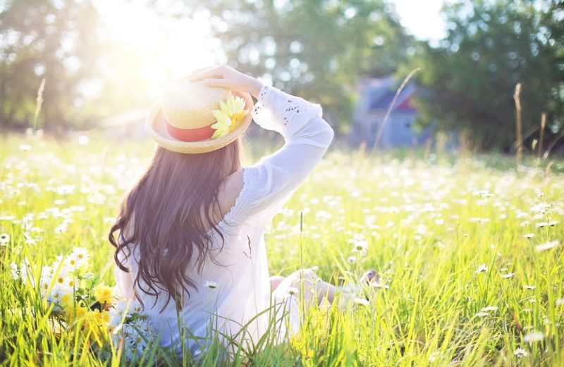 Natural ways to prevent skin cancer: Image of young woman, shot from behind, sitting on ground in sunny field with wide brimmed hat on.