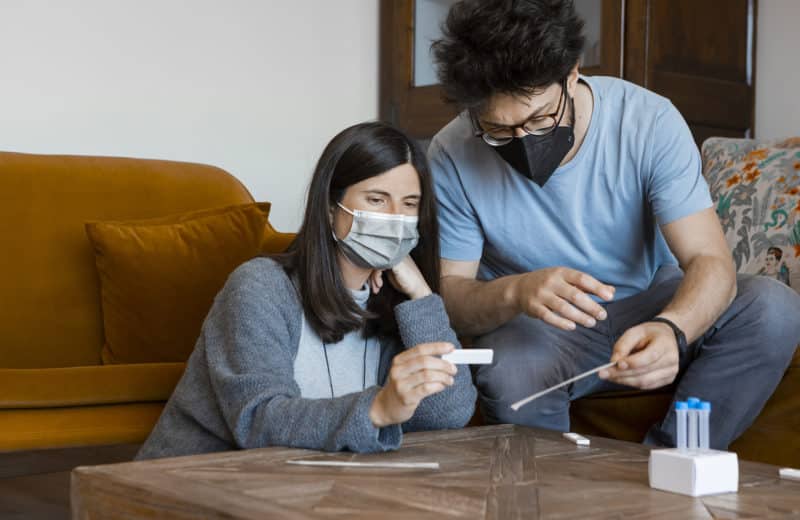 Man and woman in their 30s, wearing surgical face masks, sitting in the living room at home, checking the results of the self-swabbing Antigen Home Test for Coronavirus diagnostic.