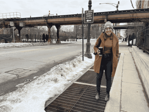 Heather Bodie stands outside in a long brown coat, holding a coffee on a Chicago street. L tracks are in the distance behind her.