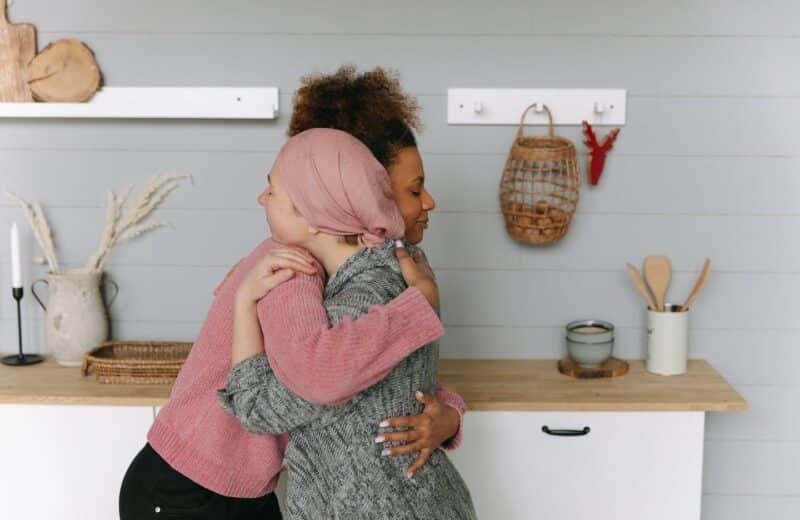 Two women hugging in a kitchen, and one has colorectal cancer. One of the women has a shaved head and a pink wrap covering.