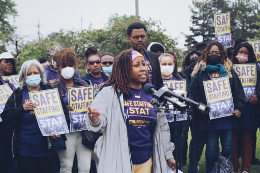 Healthcare worker in a purple shirt and gray cardigan speaks about staffing shortages at hospitals, standing before a group of people holding signs that say "Safe Staffing Stat"