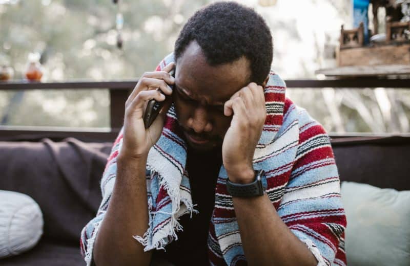 Black man has a striped red, white, and blue blanket around his shoulders. He's sitting and on a phone call, looking upset.