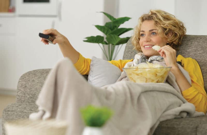 Woman sitting on sofa snacking with remote control demonstrating a sedentary lifestyle