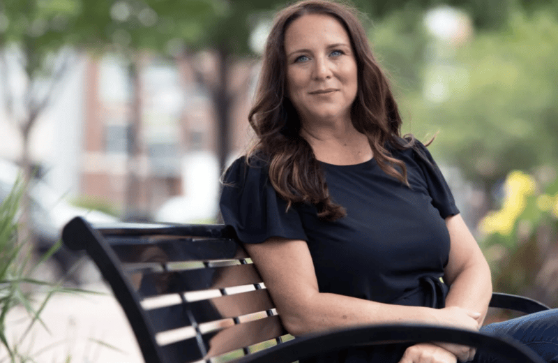 Cathy Cassata, with shoulder-length brown hair, sits on a park bench in the summer wearing a navy blue t-shirt and jeans, looking at the camera.