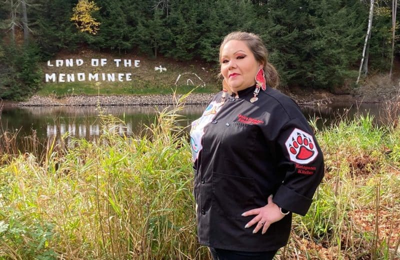 Chef Jessica Walks First stands in her black chef's shirt and pants beside a lake. Land of the Menominee is written in white on a hill in the distance.