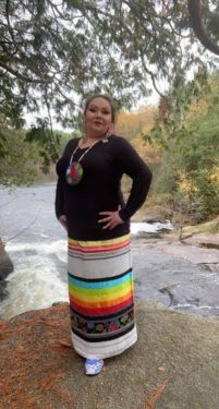 Chef Walks First stands by a river in a traditional skirt with brightly colored stripes, black shirt, and large red traditional necklace