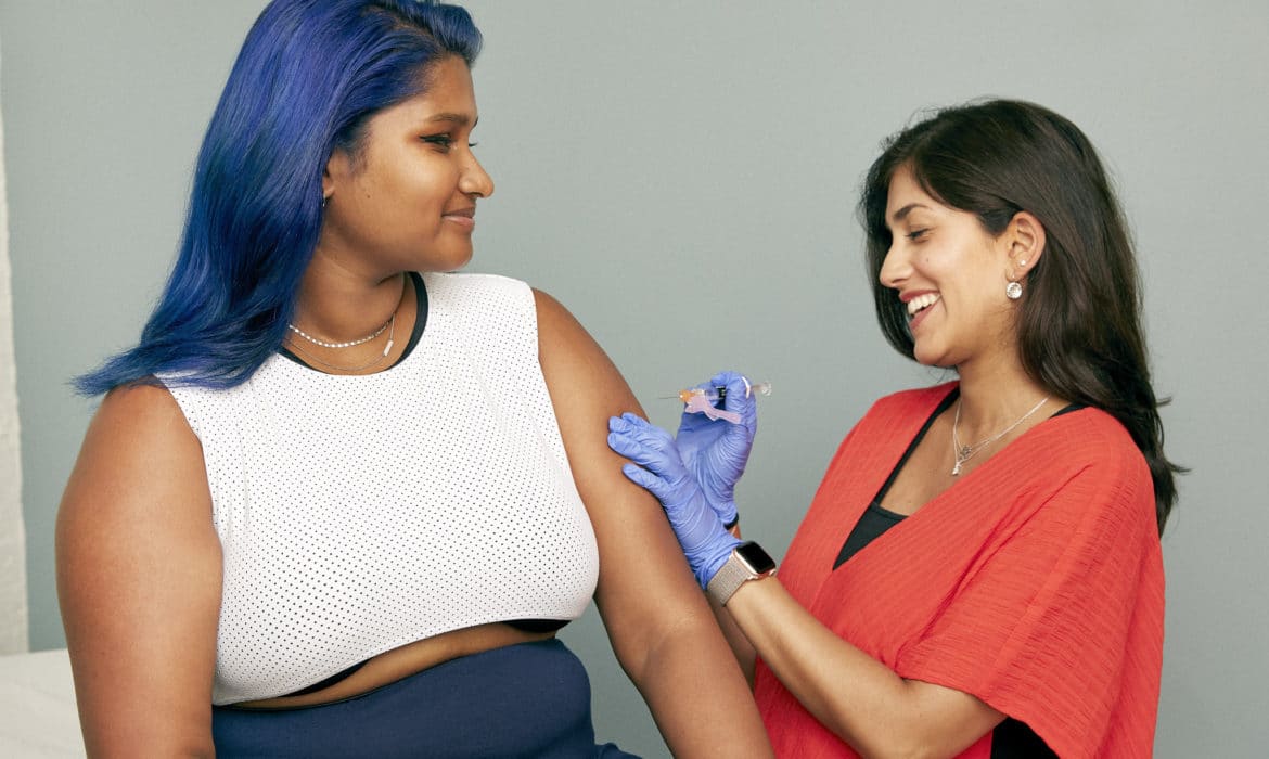 Young woman in a white crop top with blue hair about to receive a vaccine from a clinician in orange scrubs, wearing blue gloves.