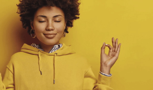 A Black woman in a yellow sweatshirt stands against a yellow background. Her eyes are closed, and she has a slight smile on her face. She's holding her fingers in a meditative posture with index fingers lightly touching.