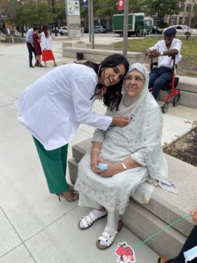 A medical student at a White Coat Ceremony stands next to her grandmother, with a stethoscope to the grandmother's heart. They're smiling, looking at the camera.