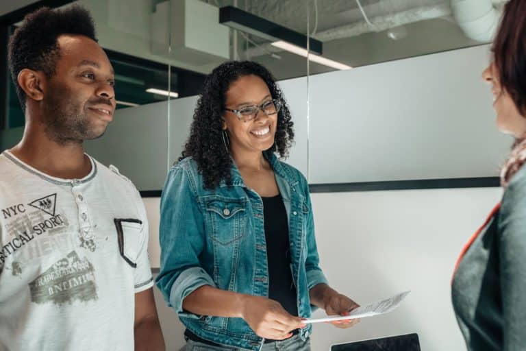 A teenager and his mom stand beside each other smiling at a lawyer in an office. The mom has glasses and is holding paperwork.