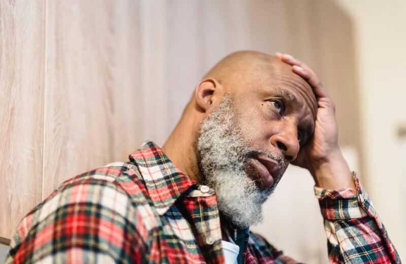 Bald man with beard, in red, white, and black plaid shirt, holds hand to head, looks deep in thought.