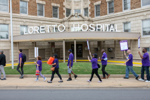 Strikers outside of Loretto Hospital march in a line, holding signs and wearing purple shirts.