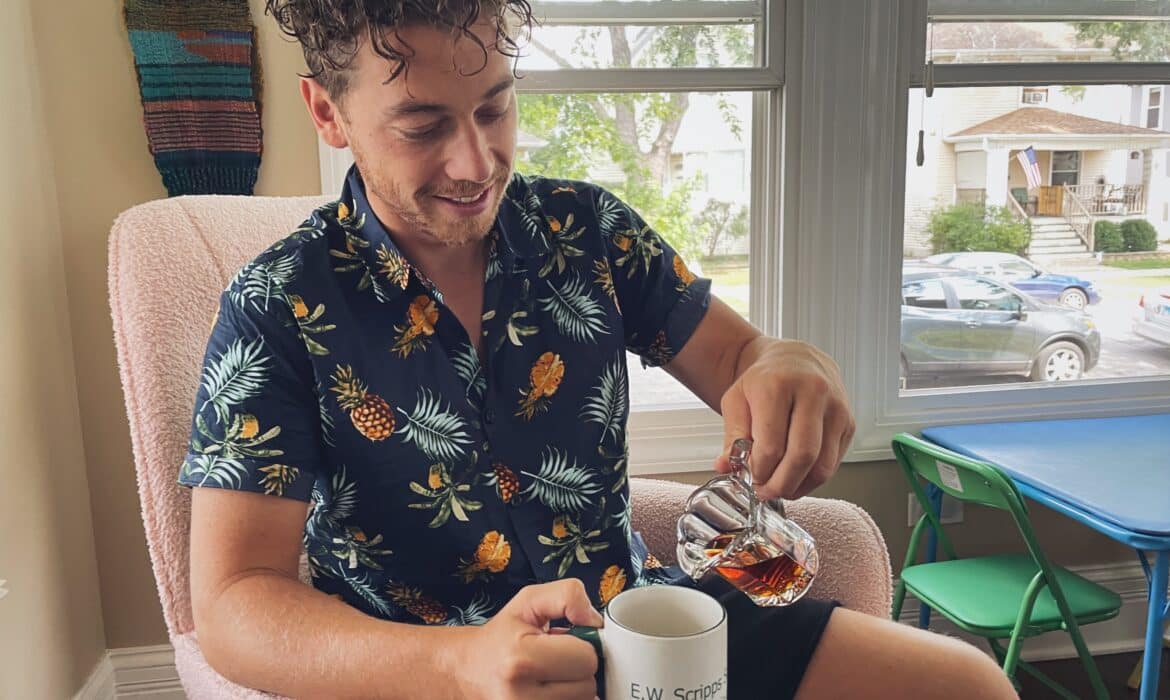 A curly haired man sits in a pink rocking chair alongside a series of open windows. He's wearing a button up flowered shirt, pouring maple syrup from a glass pitcher into a white coffee mug.