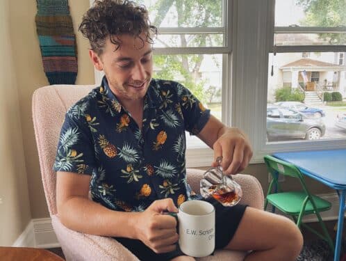 A curly haired man sits in a pink rocking chair alongside a series of open windows. He's wearing a button up flowered shirt, pouring maple syrup from a glass pitcher into a white coffee mug.