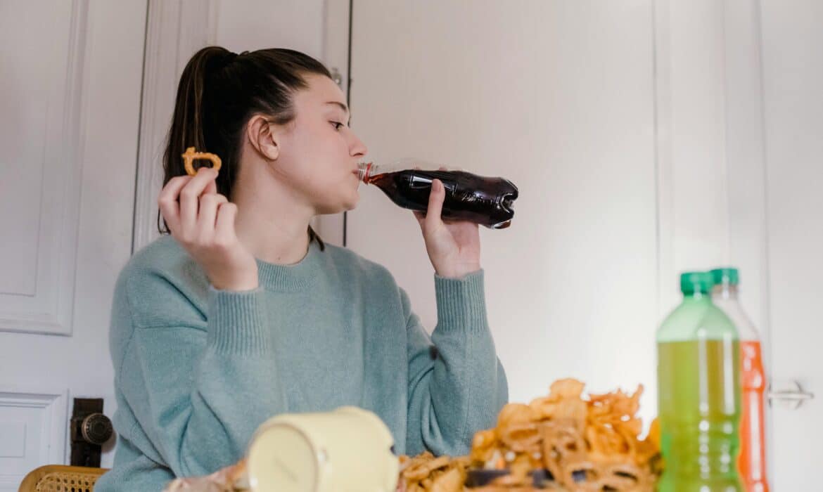 Woman at a table, wearing a blue sweatshirt and ponytail, is drinking soda and sitting in front of a pile of snacks.