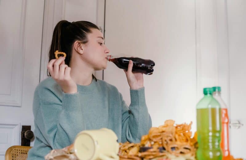 Woman at a table, wearing a blue sweatshirt and ponytail, is drinking soda and sitting in front of a pile of snacks.