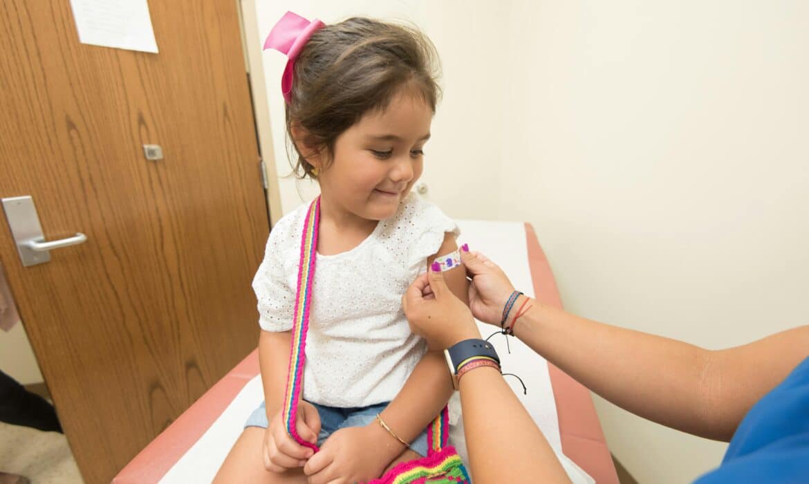 Young girl with brown hair and a pink bow gets vaccinated in a patient room. She is wearing a white tshirt and watching the practitioner give the shot.