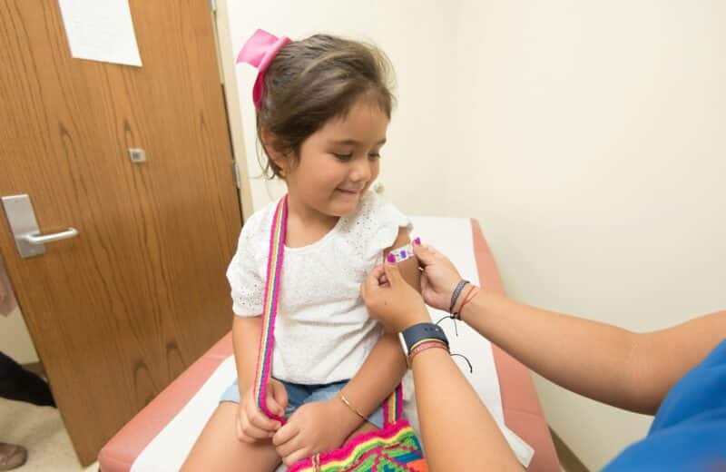 Young girl with brown hair and a pink bow gets vaccinated in a patient room. She is wearing a white tshirt and watching the practitioner give the shot.