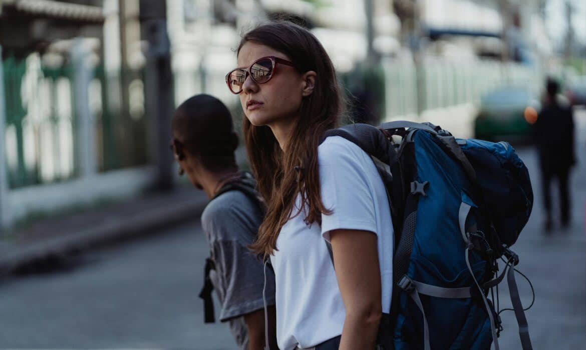 A woman in a white tshirt and sunglasses looks into the distance. She's in an urban setting, wearing a blue backpack.
