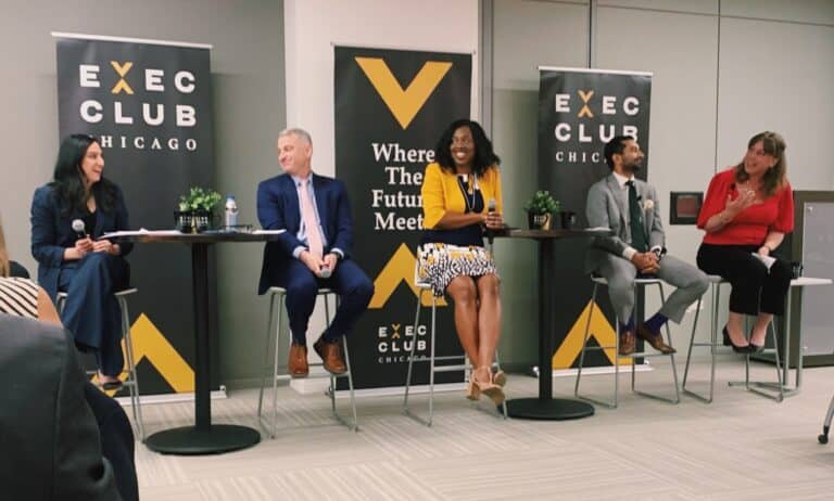 Panelists sit in tall chairs on a stage before Exec Club Chicago signage. The four panelists and moderator are smiling and laughing.