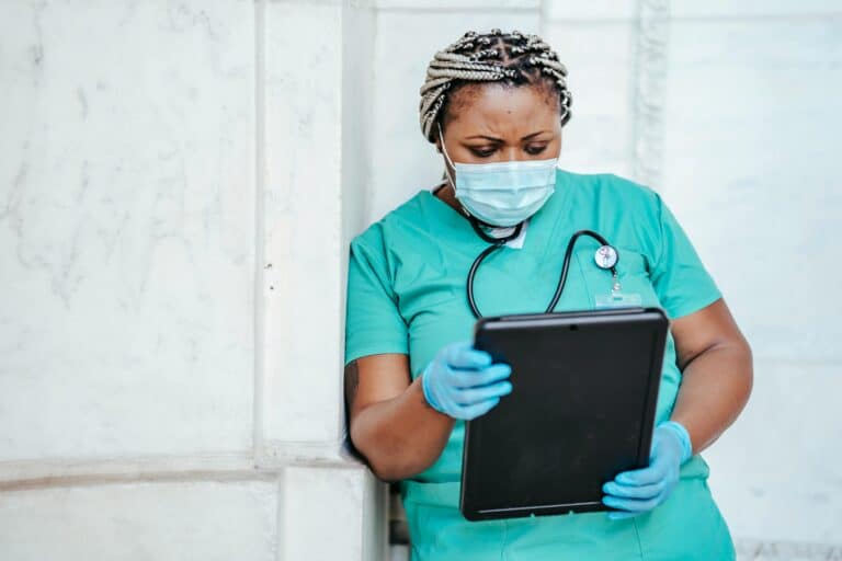 A nurse in green scrubs and a blue surgical mask leans against a building wall, looking at a tablet.