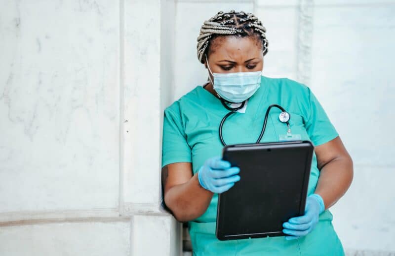 A nurse in green scrubs and a blue surgical mask leans against a building wall, looking at a tablet.