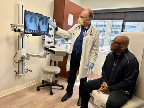 A physician points to x-rays while a patient sits on a chair and listens.