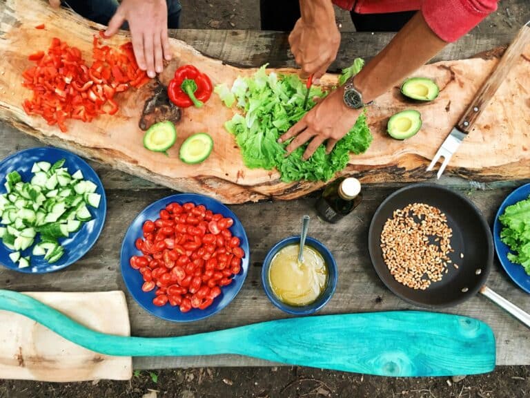 People chop up vegetables in a rainbow of colors, from red tomatoes to green lettuce. Bowls of brown and white seeds also line the cutting board. Cooking for the climate