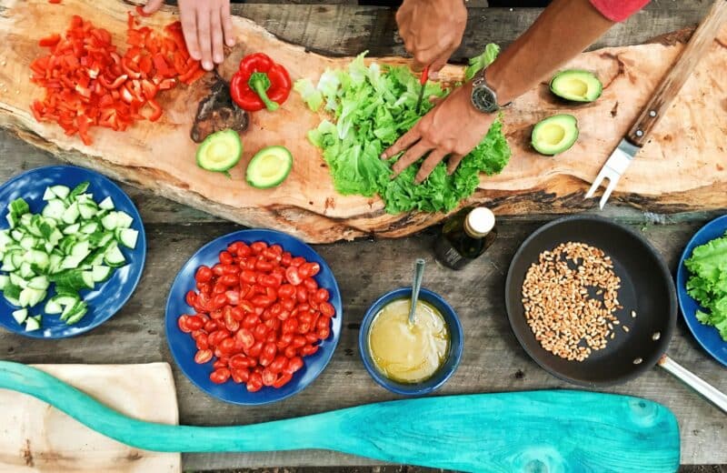 People chop up vegetables in a rainbow of colors, from red tomatoes to green lettuce. Bowls of brown and white seeds also line the cutting board. Cooking for the climate