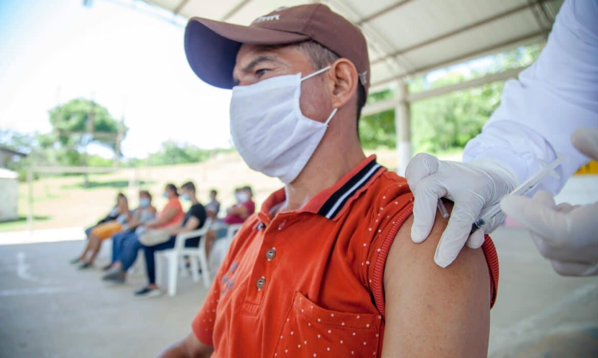 A man in a red tank top, white surgical mask, and brown baseball hat stares into the distance as a gloved hand gives him a vaccination.