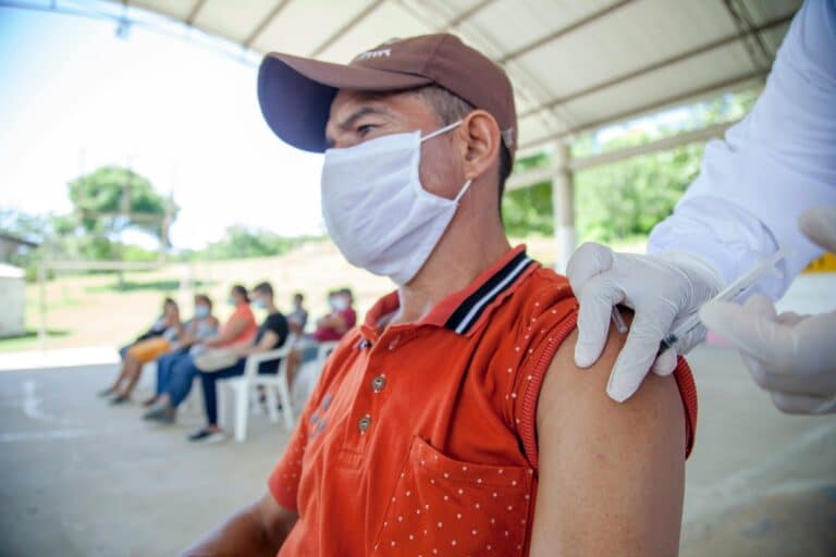 A man in a red tank top, white surgical mask, and brown baseball hat stares into the distance as a gloved hand gives him a vaccination.