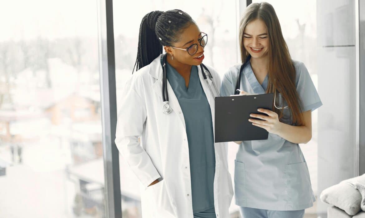 A female physician talks with a nurse; they're standing in front of large windows, looking at an ipad and smiling