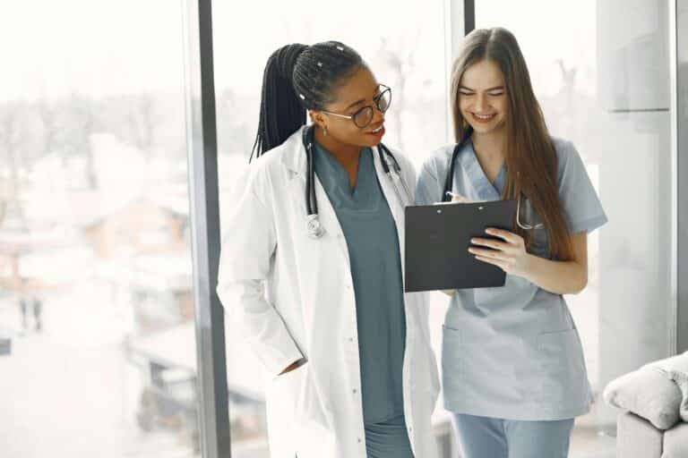 A female physician talks with a nurse; they're standing in front of large windows, looking at an ipad and smiling