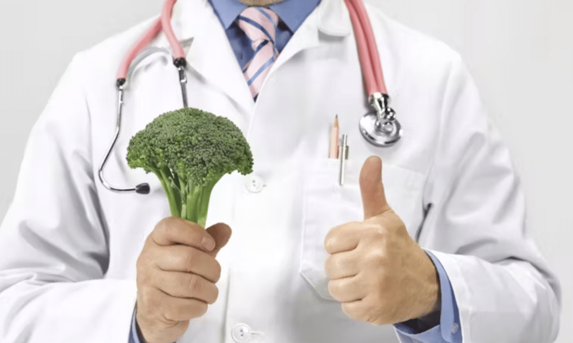 A man in a white coat stethoscope around his neck, holds a broccoli crown and gives a thumbs up sign.