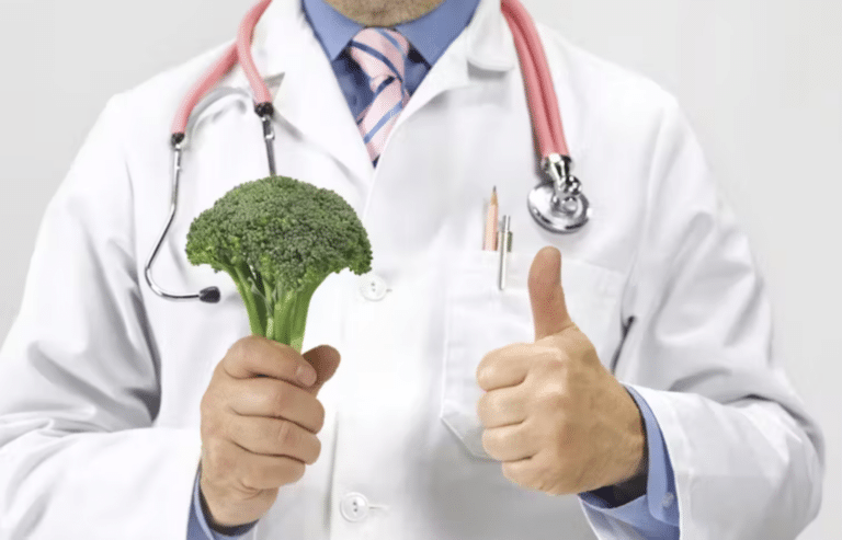 A man in a white coat stethoscope around his neck, holds a broccoli crown and gives a thumbs up sign.