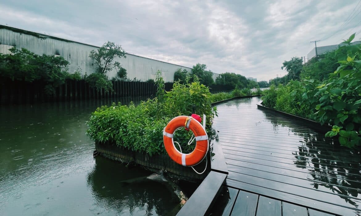 At the Wild Mile, along the Chicago River, in Chicago, facing west. A bright orange life preserver is at the center of the image, which shows the river walk on a rainy day. A white, industrial building stands in the background.