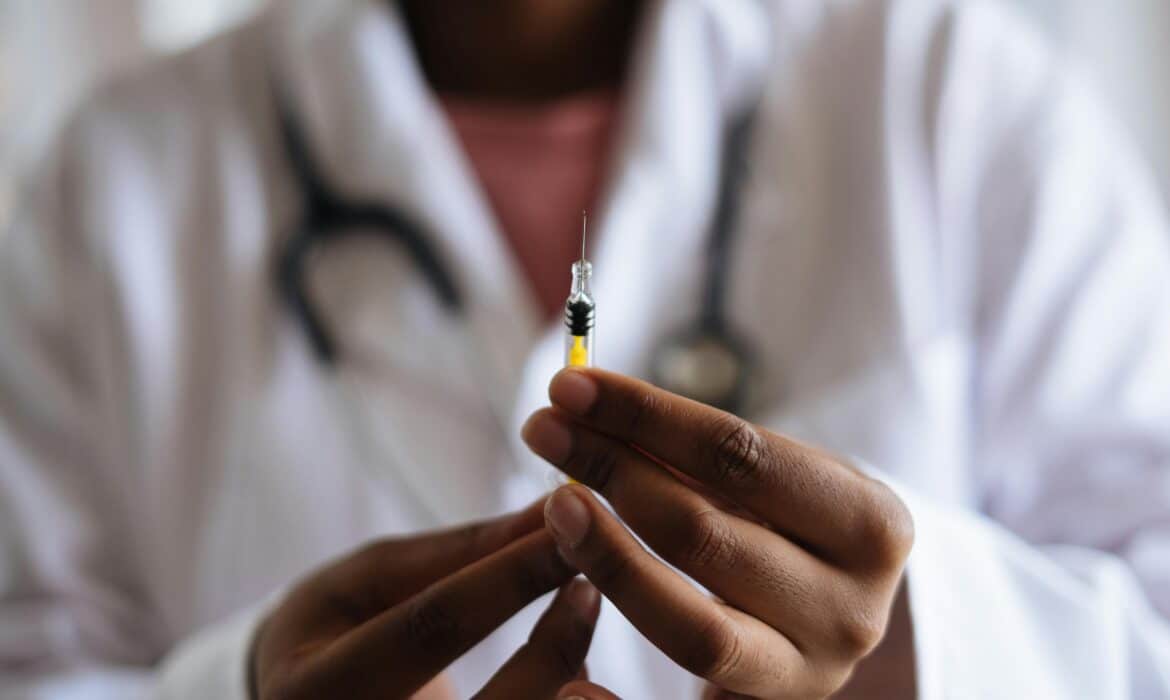 Close up of a vaccine needle with a doctor holding it and blurred in the background. The doctor is wearing a white coat and stethescope