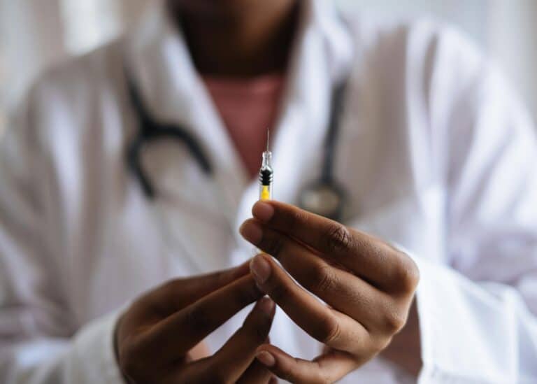 Close up of a vaccine needle with a doctor holding it and blurred in the background. The doctor is wearing a white coat and stethescope