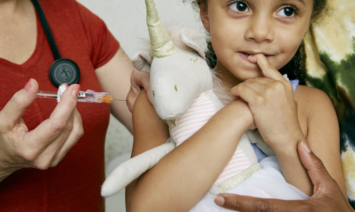 A little girl snuggling a unicorn looks brave but nervous before receiving a vaccination.