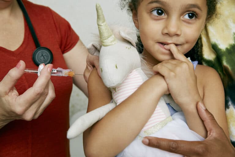A little girl snuggling a unicorn looks brave but nervous before receiving a vaccination.