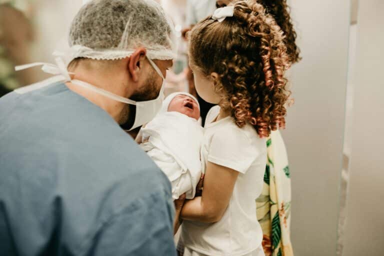 A man in scrubs and a surgical mask holds a new baby for his daughter to see.
