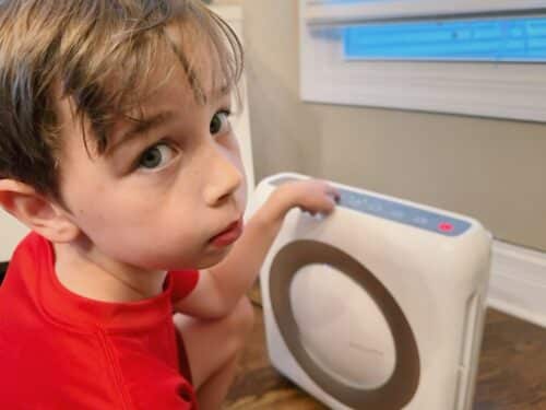 A young child looks toward the camera forlornly, as he presses the button to turn on the home's air purifier. The purifier's light is red, indicating polluted air in the home.