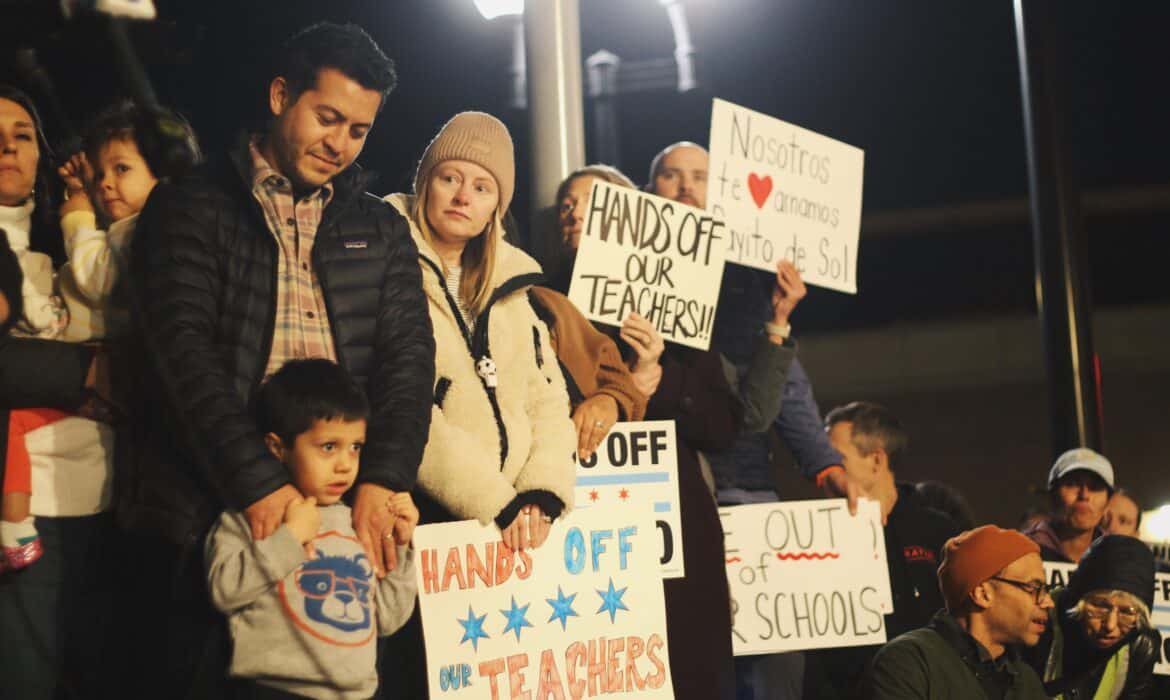 Families stand on stage holding signs during a rally for Rayito de Sol teacher Ms. Diana