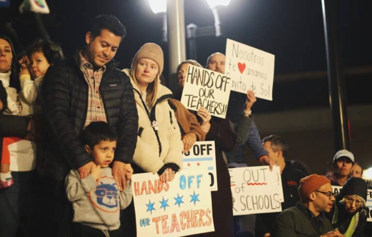 Families stand on stage holding signs during a rally for Rayito de Sol teacher Ms. Diana