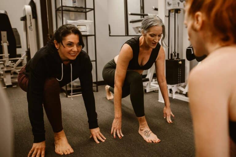 Two women in workout clothes stretch in a lunge formation at a gym.