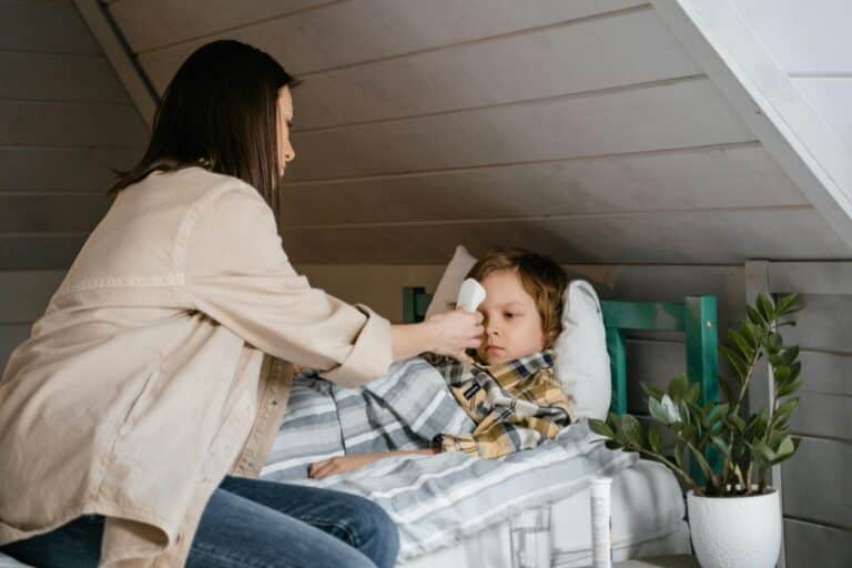 A mom sits at the edge of her son's bed, taking his temperature.